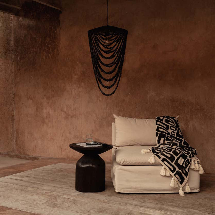 Beige sofa with a patterned blanket in a room with a textured brown wall and black beaded chandelier.