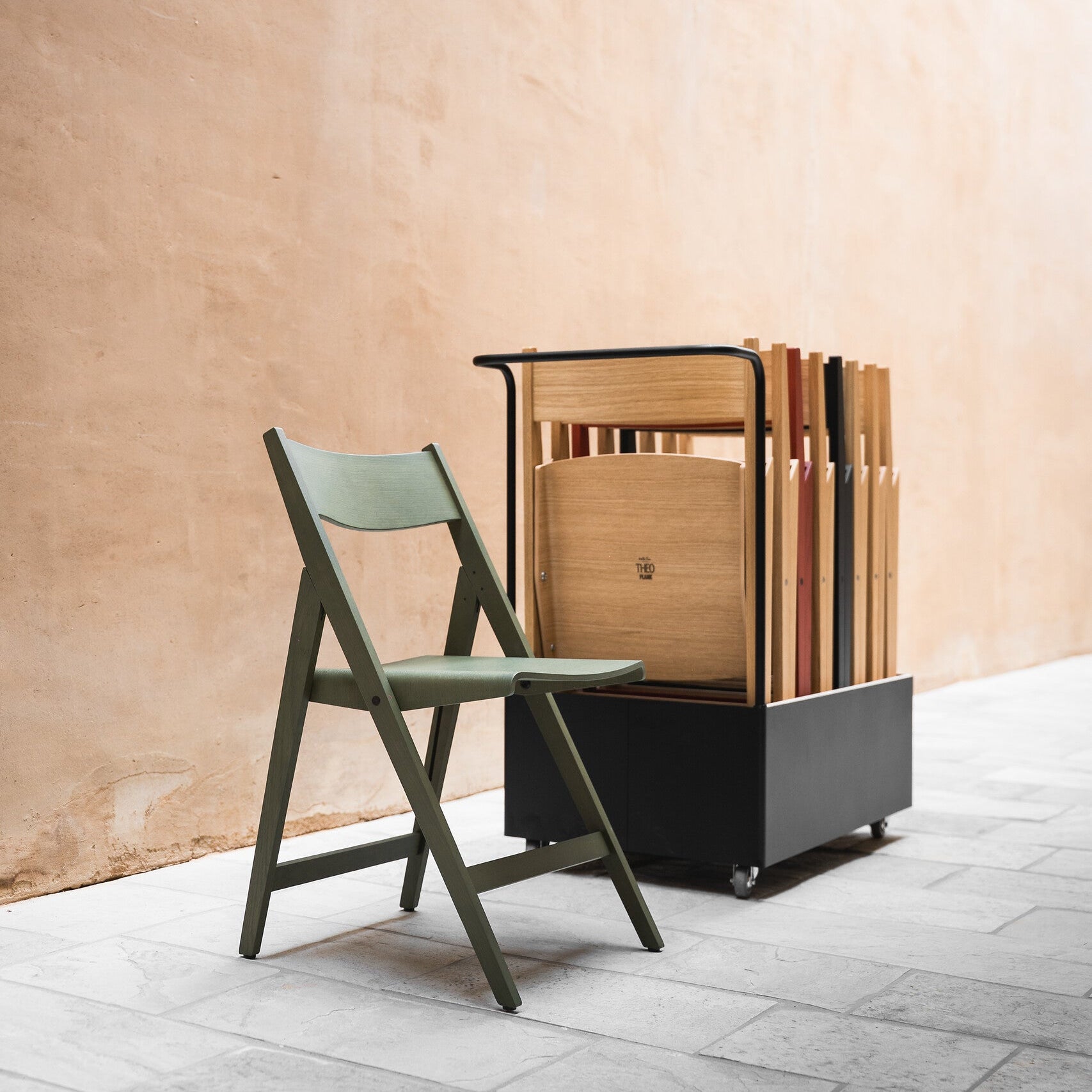 Green THEO folding chair next to a black trolley with stacked chair against a beige wall.